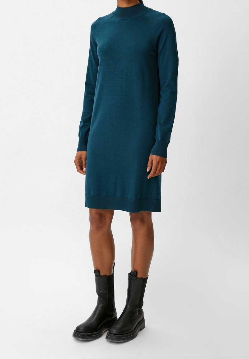Abito lavorato a maglia teal con collo alto e maniche lunghe, caratterizzato da un orlo a coste. Abbinato a stivali neri alla caviglia con suola spessa.