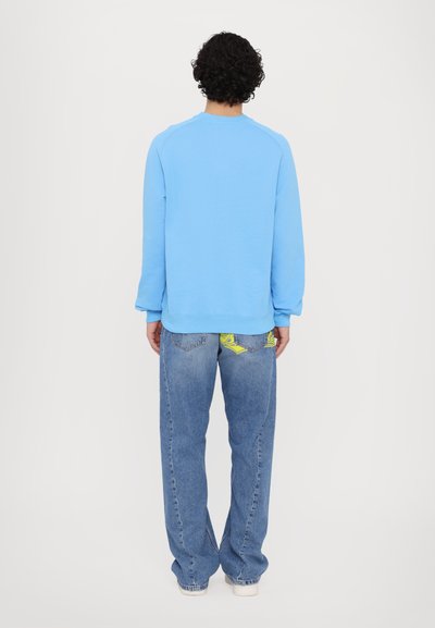 Sudadera azul clara con un cuello redondeado y puños de canalé, combinada con unos pantalones vaqueros azules de talle alto y corte holgado con detalles amarillos.