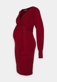 Robe de maternité tricotée rouge avec des manches longues, un col en V et une ceinture à la taille, conçue pour accueillir un ventre de grossesse.