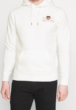 Homme portant un sweat à capuche blanc avec poche avant et logo sur la poitrine indiquant « GANT 1949 American Sportswear » et un pantalon noir.