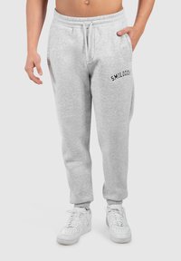 Jasnoszare dresowe spodnie z elastycznym pasem, sznurkiem, bocznymi kieszeniami i czarnym logo "SMILODOX" na lewej udzie. Białe sneakersy.