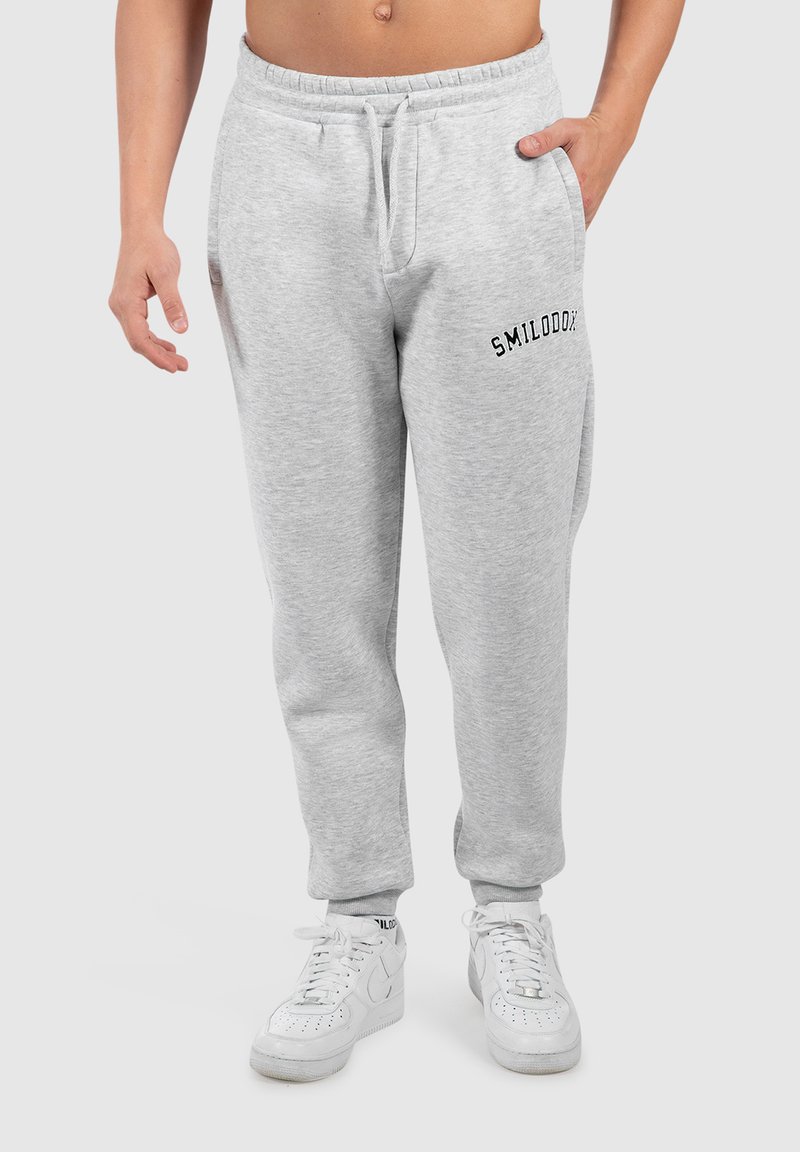 Jasnoszare dresowe spodnie z elastycznym pasem, sznurkiem, bocznymi kieszeniami i czarnym logo "SMILODOX" na lewej udzie. Białe sneakersy.