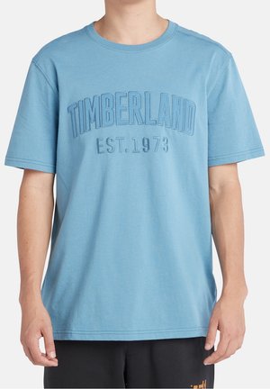 Vaaleansininen lyhythihainen t-paita, jossa on kohotettu "TIMBERLAND EST. 1973" -teksti, käytössä valkoista taustaa vasten.