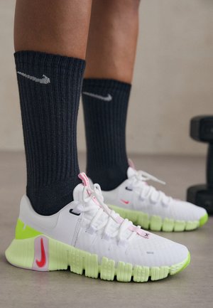 Baskets Nike blanches et vert néon portées avec des chaussettes Nike noires, debout sur un sol lisse près d'un haltère.