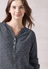 Blusa a motivi bianco e nero con linee geometriche, scollo a V e dettagli con bottoni nella parte frontale. Maniche lunghe con una vestibilità rilassata.