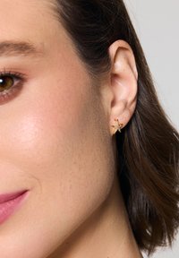 Złoty postrzępiony w kształcie rozgwiazdy kolczyk z teksturalnymi detalami i małymi kamieniami szlachetnymi, noszony na płatku ucha, komplementujący krótkie, ciemne włosy oraz gładką skórę.