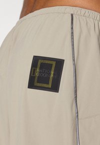 Beige shorts med elastisk linning, en slank svart og gul logo-patch, og subtile sidepaneldetaljer.