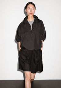 Giacca nera a pullover con colletto a mezza cerniera, abbinata a una gonna nera con tasche drappeggiate. Entrambi i capi hanno una texture liscia.