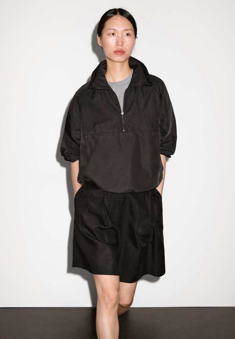 Giacca nera a pullover con colletto a mezza cerniera, abbinata a una gonna nera con tasche drappeggiate. Entrambi i capi hanno una texture liscia.