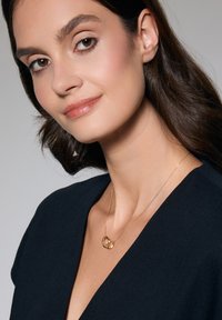 Złoty naszyjnik z przeplatającymi się pierścieniami, delikatny łańcuszek, noszony na ciemnej bluzce z dekoltem w serek, podkreślający design i formę biżuterii.