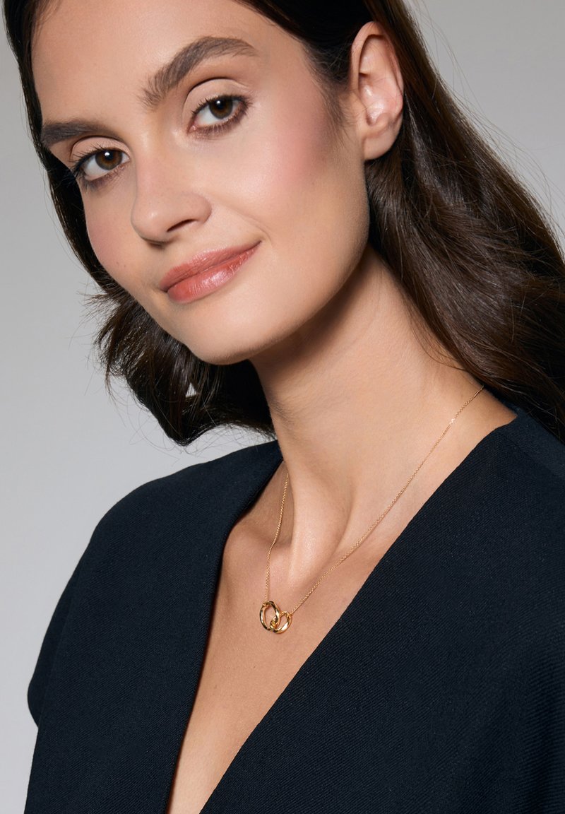 Złoty naszyjnik z przeplatającymi się pierścieniami, delikatny łańcuszek, noszony na ciemnej bluzce z dekoltem w serek, podkreślający design i formę biżuterii.
