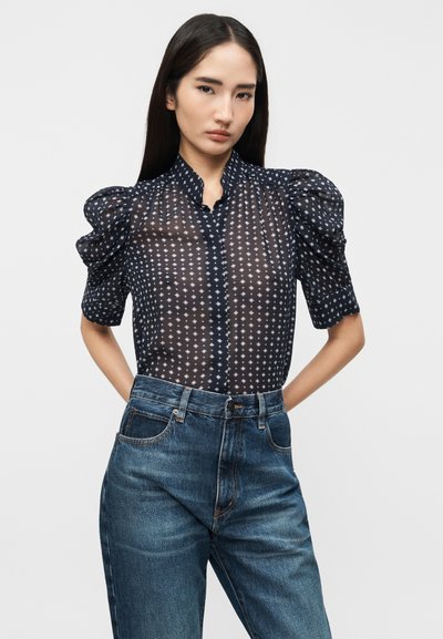 Γυναίκα με μακριά σκούρα μαλλιά που φοράει διαφανή μπλούζα σε ναυτικό μπλε με φουσκωτά μανίκια και λευκά διαμάντια σχεδίου, συνδυασμένη με ψηλόμεσο μπλε τζιν.