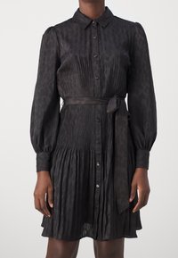 Vestido negro de tela suave con cuello, botones en la parte delantera, mangas largas, pliegues y una cinturilla atada. Detalle sutil de patrón de leopardo.