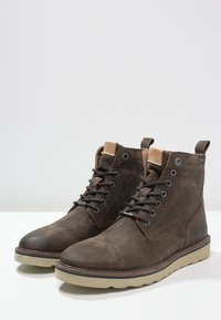 Stivali ankle in suede marrone con lacci, colletto imbottito beige e suole in gomma beige chiaro su una superficie bianca.