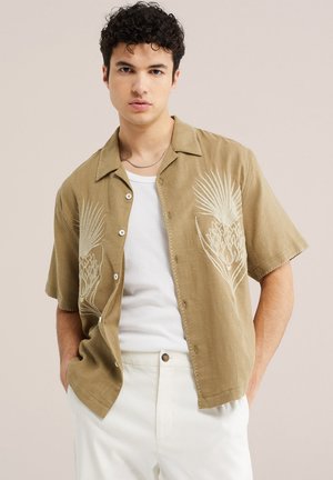 Jeune homme aux cheveux bouclés portant une chemise beige à manches courtes avec des impressions botaniques blanches par-dessus un T-shirt blanc et un pantalon de couleur claire.