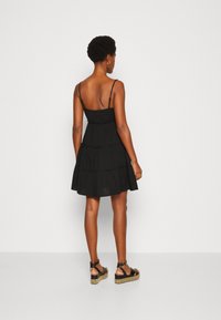 Svart, lager-på-lager sommarklänning med tunna remmar, tillverkad av lättviktsmaterial. Har en åtsittande bodice och en flödande kjol, samt styva svarta kilsandaler.