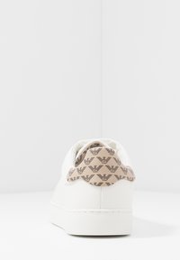 Vita sneaker med beige hälla som har ett repetitivt mönster av svart logotyp, visad från en bakre vy på en vit yta och bakgrund.