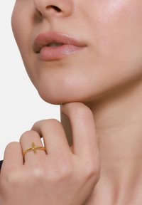 Bague en métal doré avec un design en croix, surface lisse, bande fine, portée sur une main avec une couleur d'ongles neutre, mettant en valeur la texture naturelle de la peau.