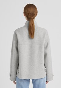 Graue Wollmischungjacke mit hohem Kragen, fallenden Schultern und Knopfdetails an den Ärmelbündchen, die einen entspannten, übergroßen Schnitt hat.