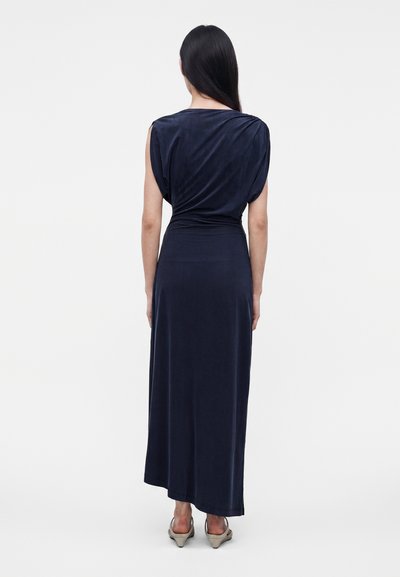 Femme aux longs cheveux lisses vue de dos, portant une robe bleu marine sans manches jusqu'aux chevilles et des talons beiges sur un fond uni.
