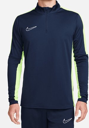 Hombre vistiendo una camiseta deportiva de manga larga azul marino y amarillo neón con cremallera y pantalones a juego, con logotipos de Nike visibles en la camiseta y los pantalones.