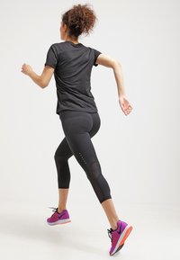 Camiseta atlética negra, leggings capri negras con paneles de malla y zapatillas de correr moradas con suelas grises y detalles en naranja, en una postura de carrera.