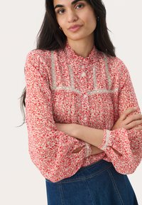 Blusa rossa floreale con colletto arricciato, dettagli in pizzo e chiusura a bottoni. Le maniche lunghe presentano ritagli con rifiniture in pizzo. Indossata sopra una gonna di jeans.