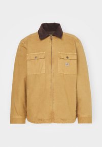 Tan jacka med ett kordflätskrage, frontdragkedja och två bröstfickor. Har detaljerade sömmar och ett Lee-logotypmärke på vänster ficka.