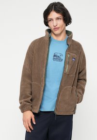 Jauns vīrietis valkā brūnu zip-up flīsa jaku pār zilu grafisku t-kreklu un tumšām biksēm uz vienkārša fona.