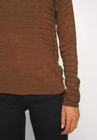 Personne portant un pull en maille marron à manches longues et un pantalon noir, visible de la moitié du torse à la main sur un fond blanc uni.