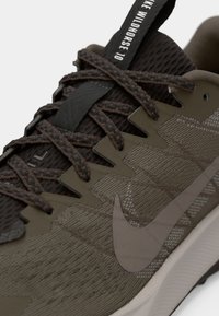 Tuvumā Brown un Olīvu krāsas Nike Wildhorse 10 sneakers, parādot strukturālu audumu, šņorītes un lielu swoosh logotipu uz zoles maliņas.
