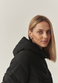 Crna puffer jakna s kapuljačom, koja ima kvadratni dizajn i kopčanje na dugmad. Model ima ravnu plavu kosu i nosi male zlatne naušnice.