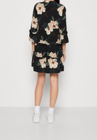 Vestido negro con flores beiges, presenta un diseño en capas y mangas tres cuartos. Combinado con zapatillas blancas y calcetines de canalé.