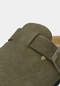 Nahaufnahme einer olivengr�ünen Wildleder-Sandale mit einem breiten Riemen und einer mattschwarzen Schnalle auf einer beigen Fußbett.