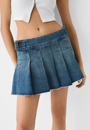 Haut court blanc côtelé avec fermeture éclair sur le devant, associé à une mini-jupe plissée en denim bleu taille haute avec détail à double boucle.