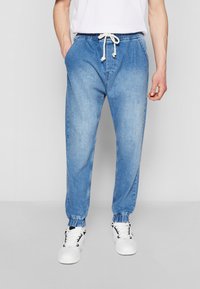 Vaaleansiniset denim-jogger-housut, joissa on joustavat hihnat nilkoissa ja säädettävä nyöri vyötäröllä. Housuissa on sivutaskut ja rento malli.
