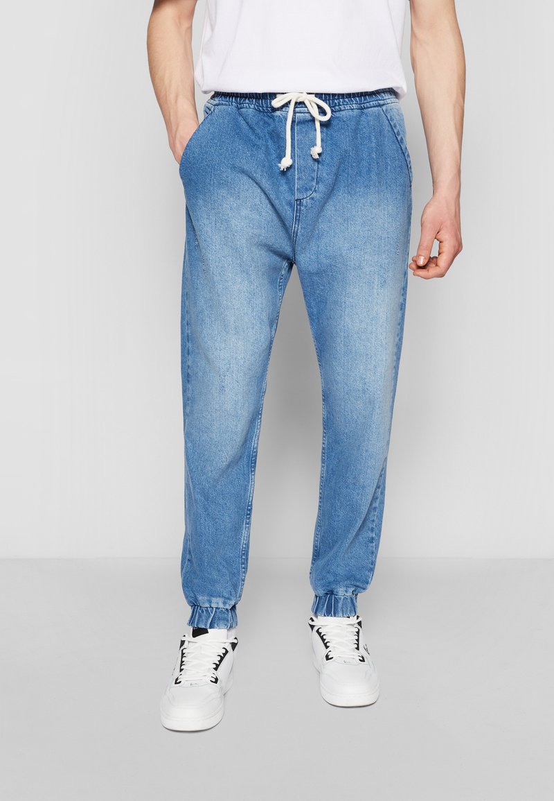 Vaaleansiniset denim-jogger-housut, joissa on joustavat hihnat nilkoissa ja säädettävä nyöri vyötäröllä. Housuissa on sivutaskut ja rento malli.