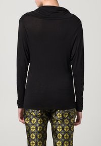Schwarzes Langarmoberteil mit einem Cowl-Neck-Design. Hergestellt aus einem weichen, dehnbaren Material. Getragen mit gemusterten gelben und grauen Hosen.