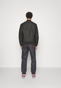 Giubbotto bomber nero con orlo e polsini a coste, abbinato a jeans in denim blu scuro con un logo visibile sulla tasca posteriore e scarpe da ginnastica.