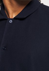 Primer plano de una camisa polo azul marino con un tejido texturado, tapeta de tres botones y un cuello suave con bordes redondeados.