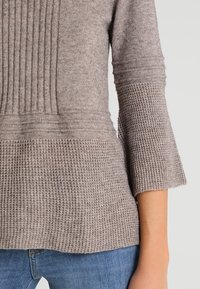 Ljust brunt, stickat tröja med ribbad textur upptill och ett våffelstickat mönster nertill, med ärmar som når till tre fjärdedelar av armarna.