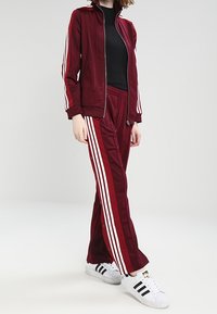 Röd träningsdräkt med vita ränder på ärmarna och sidorna av byxorna; jacka med dragkedja, byxor med elastisk midja, kombinerat med svarta och vita sneakers.