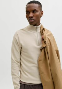 Homme portant un pull beige zippé, tenant un manteau beige sur une épaule, regardant sur le côté sur fond uni.