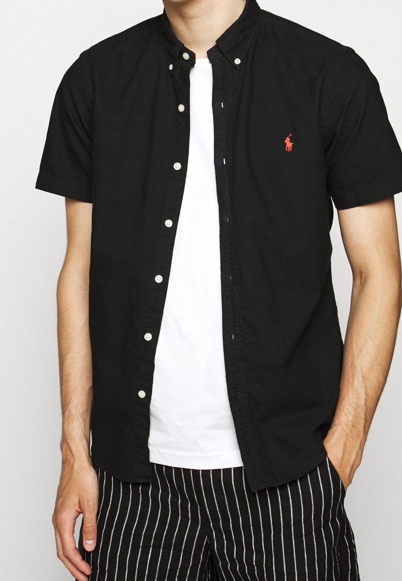 Svart kortärmad skjorta med knappar och krage, med en liten röd logotyp. Bärs över en vit t-shirt och matchas med randiga svarta byxor.