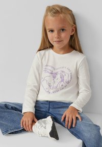 Weißes Langarmshirt mit lila Grafik von zwei Pferden und Text. Kombiniert mit blauen weiten Jeans und weißen Sneakers, im Sitzpose.