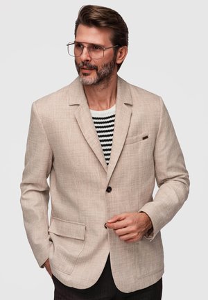 Mężczyzna z brodą i okularami ubrany w beżową marynarkę na czarno-białym paskowanym swetrze, poprawiający guzik marynarki.