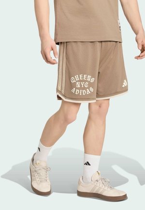 Osoba nosząca beżowe szorty z siateczki z napisem "QUEENS NYC ADIDAS", białe skarpetki Adidas i beżowe buty sportowe Adidas, stojąca.