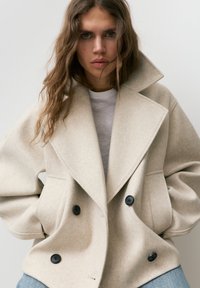 Kobieta o falowanych włosach nosi oversize beżowy dwurzędowy płaszcz z szerokimi klapami, białą koszulę i niebieskie dżinsy, patrząc prosto przed siebie.