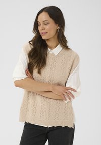 Gilet en maille beige clair avec un motif géométrique texturé, superposé sur une chemise blanche à col. Ourlet festonné et manches courtes visibles.
