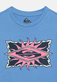 Μπλε T-shirt με στρογγυλή λαιμόκοψη, που απεικονίζει ένα ροζ και μαύρο γραφικό ηλιακής ακτινοβολίας και ένα ζωγραφιστό γράμμα "S" στο κέντρο του στήθους.